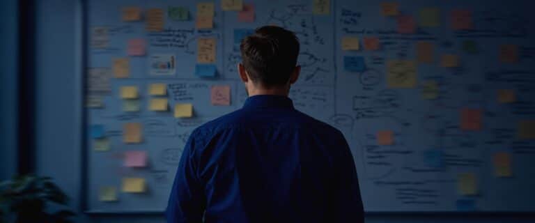 Back view of a person in a blue shirt standing before a wall covered with colorful sticky notes and drawings, suggesting brainstorming.